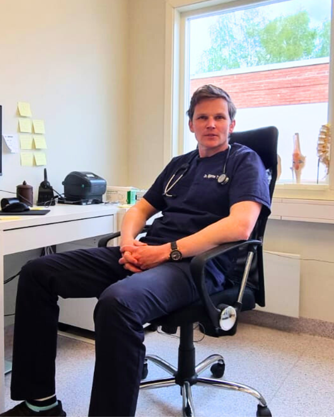 A medical doctor sitting in a chair in an office setting with a window in the background