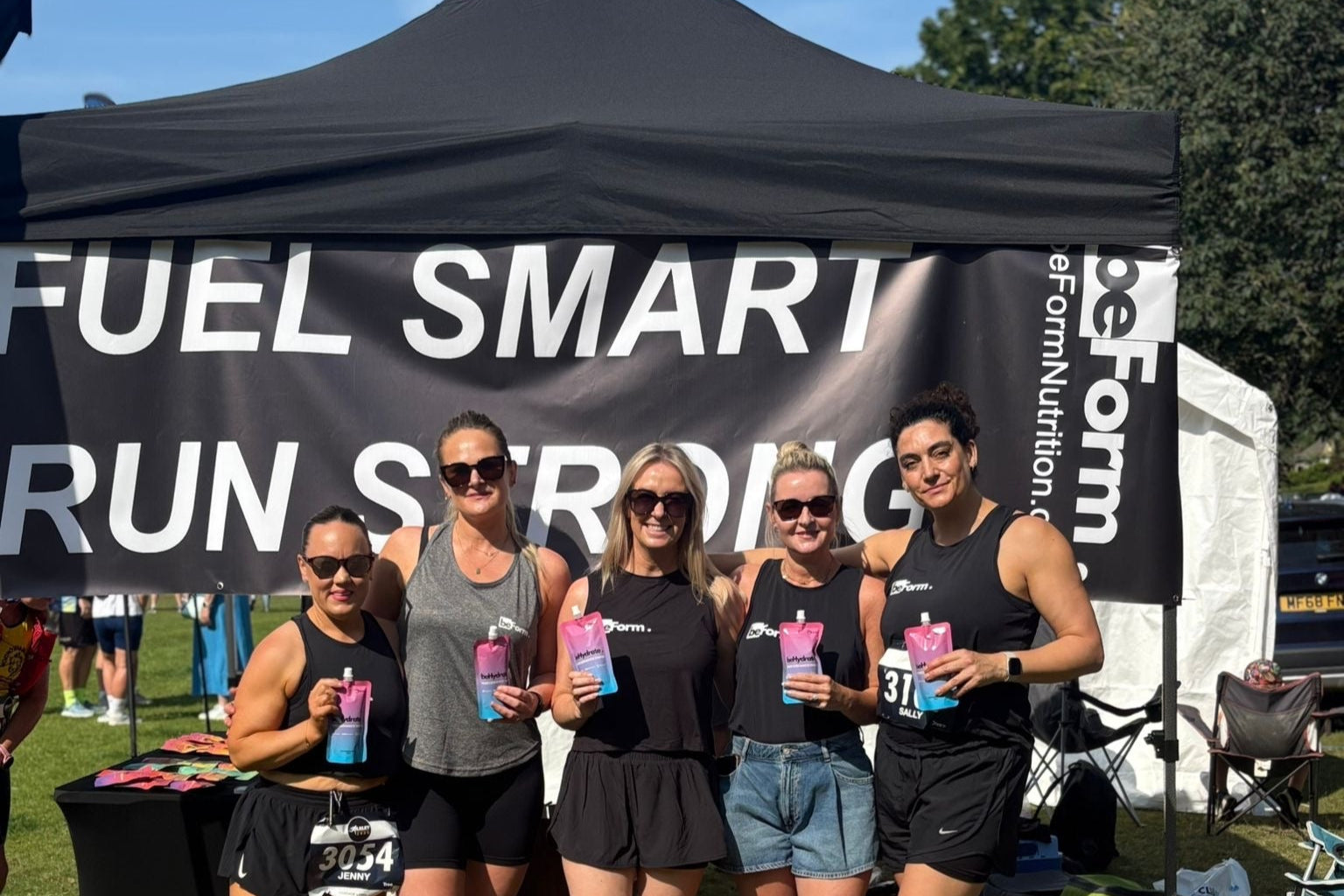 Group of runners posing in front of a 'Fuel Smart Run Strong' banner at an outdoor event holding beHydrate drink.