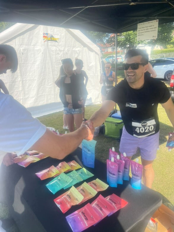 man wearing black beform tee shirt shaking hands with another person over a table of beform nutrition packets at a running event.