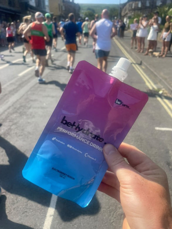 Person holding a blue and pink 'Behydrate Performance Drink' packet on a street with runners in the background