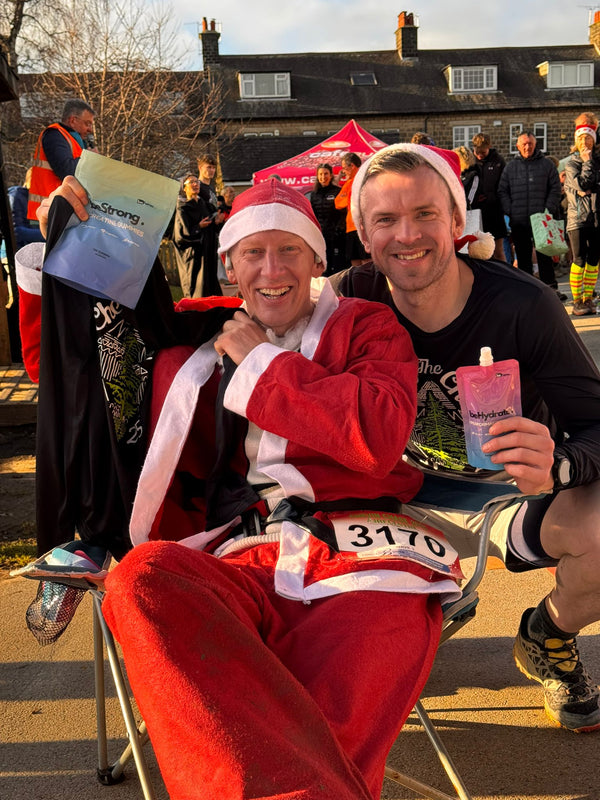 Two men, one in a Santa Claus costume and the other in casual attire, are sitting on chairs with a packet of beStrong and beHydrate at an outdoor running event.
