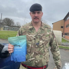 Person in military uniform holding a blue packet of beStrong creatine gummies outdoors on a cloudy day.