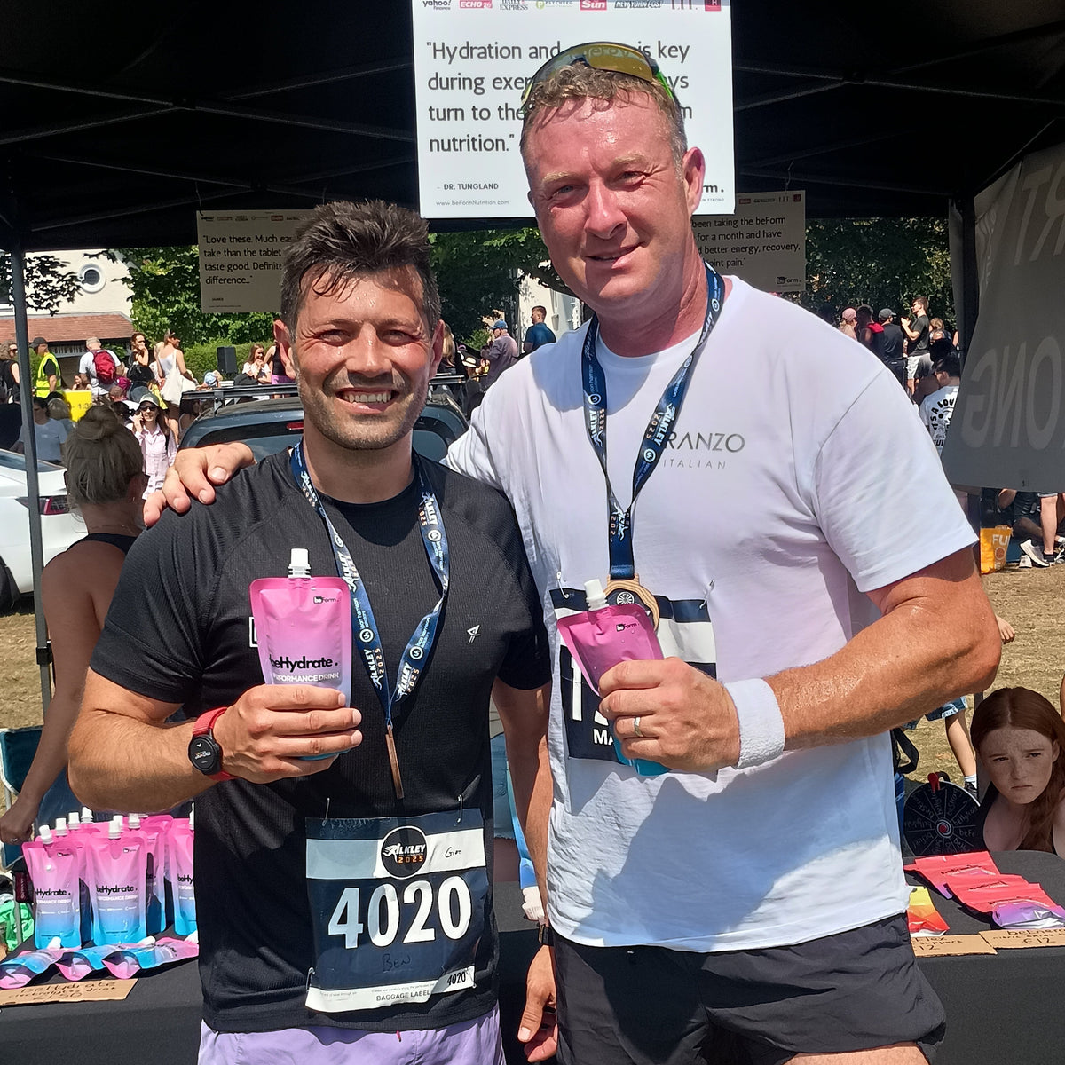Two men standing close together, one holding a pouch of beHydrate performance drink with text and the other wearing a race bib number.