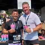 Two men standing close together, one holding a pouch of beHydrate performance drink with text and the other wearing a race bib number.