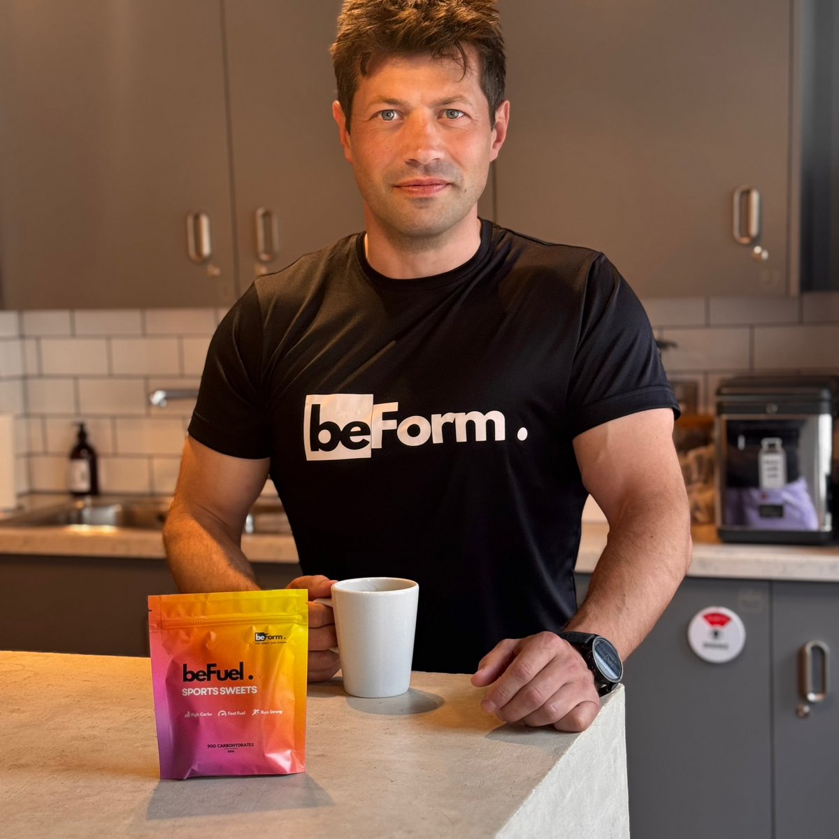 Man in a kitchen wearing a black 'beForm' t-shirt with BEfuel sports sweets on the counter.