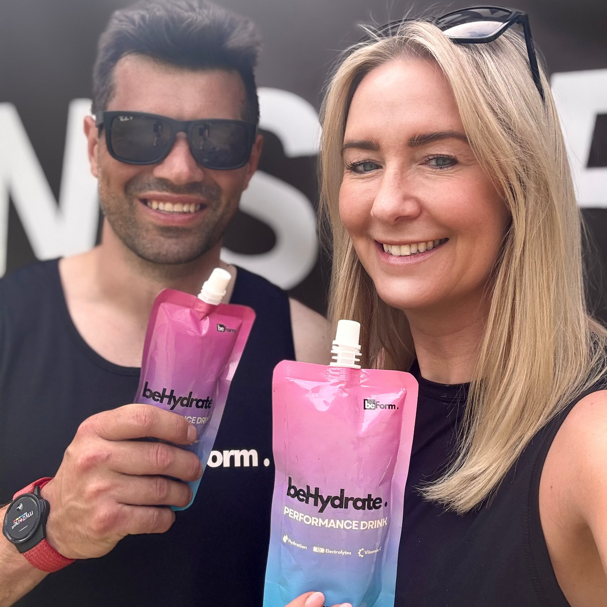 Two people holding pink 'behydrate' performance drink pouches in front of a black background with white text.
