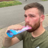 Man with brown hair and beard drinking from a pink and blue pouch of behydrate on a residential street.