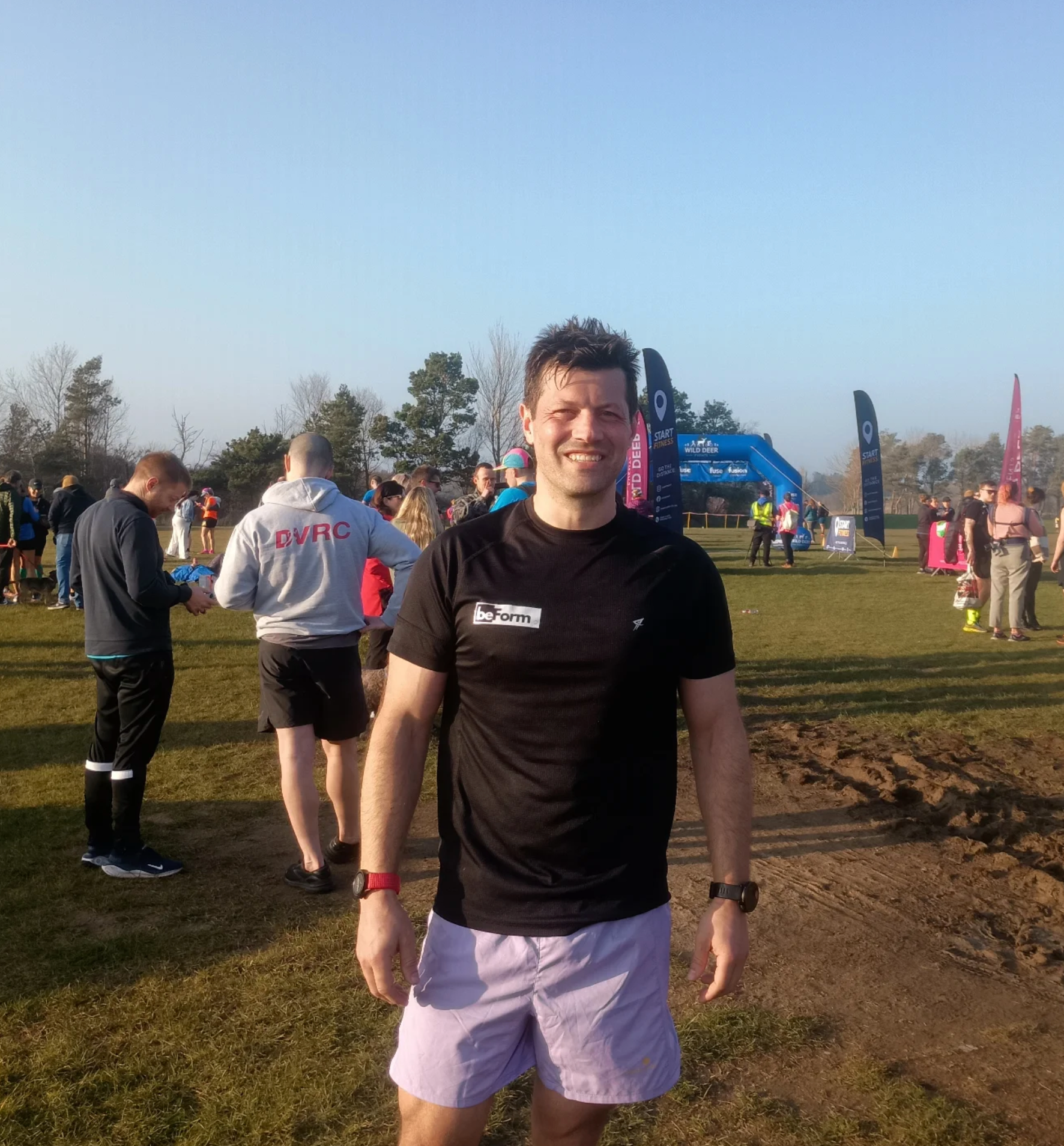 man with brown hair at a running event wearing a black tee shirt, purple shorts with the beform nutrition logo.