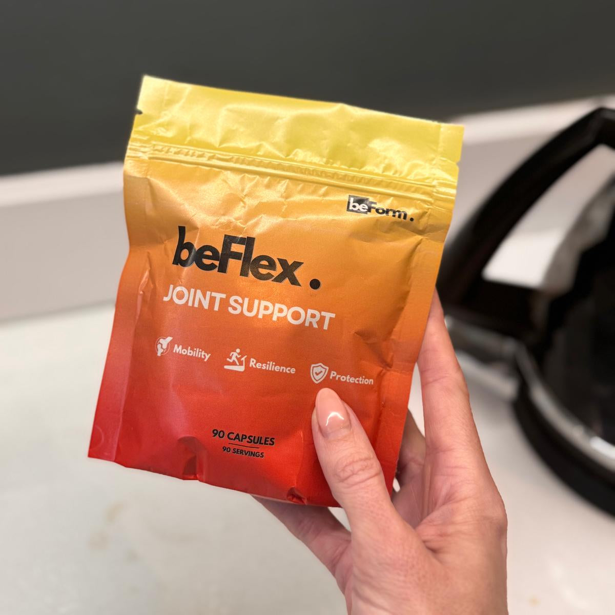 Hand holding a packet of 'beflex joint support' capsules against a neutral background.