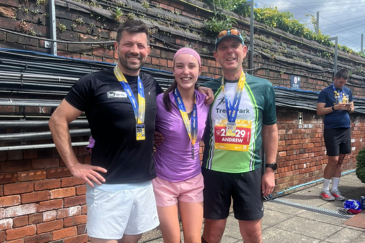 Three runners with medals standing on a street, with a brick wall and greenery in the background.