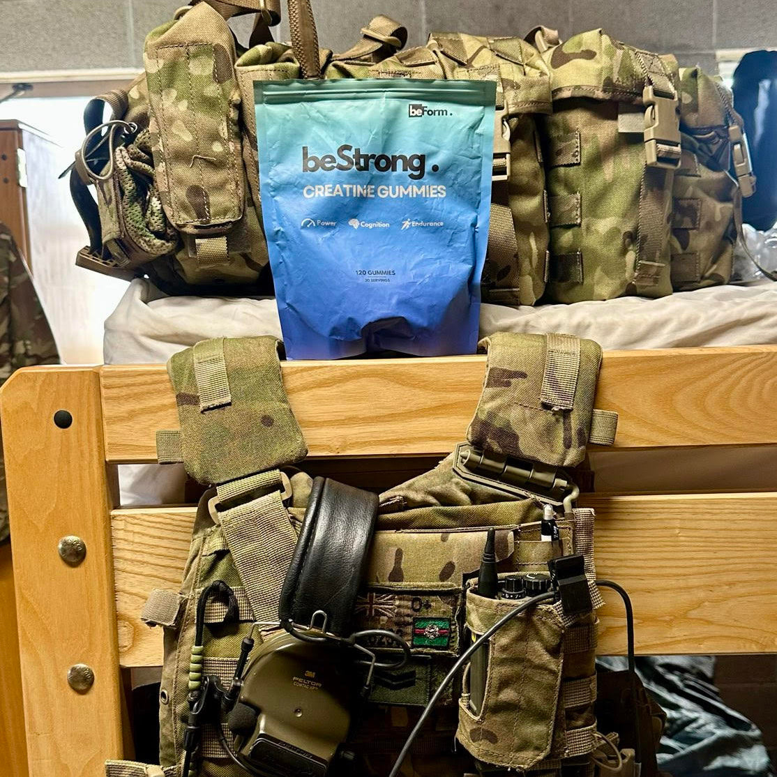 Camouflage backpack with tactical equipment and a blue 'beStrong' packet on a wooden surface.